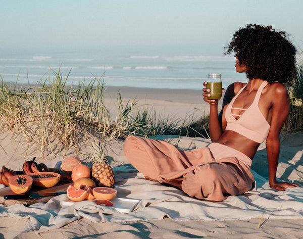 A womxn of color having a beach picnic with fresh fruits and a green juice in hand.