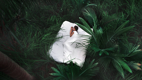 A womxn curled up in sheets on a bed, in the middle of foliage outside.