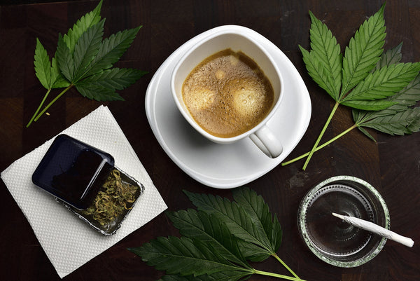 Downward shot of a cup of coffee, a pre-roll in a glass ashtray, and smokable flower in a stash jar—framed in cannabis leaves.