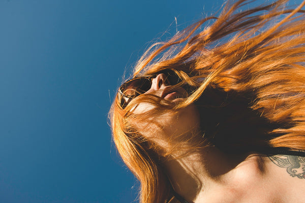 Upshot of a red-haired womxn wearing sunglasses with hair blowing in her face.