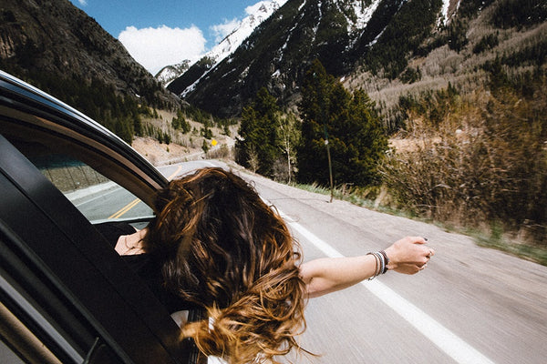 Image of the back of a womxn leaning her head and arm of a car window on a highway between tree-lined and snow-capped mountains.