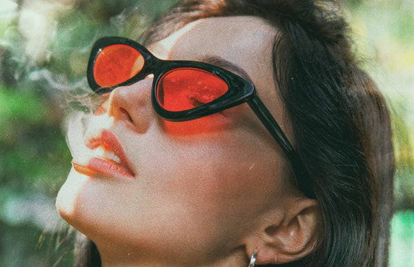 A young woman with sunglasses on, looking up while exhaling smoke.