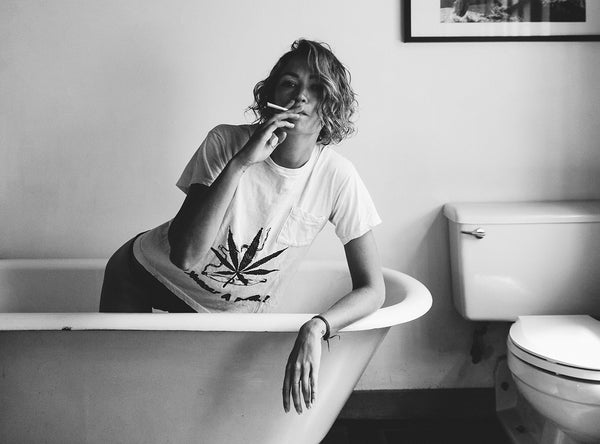 Image of woman smoking a pre-roll in a cannabis-print tee while kneeling in a bathtub.